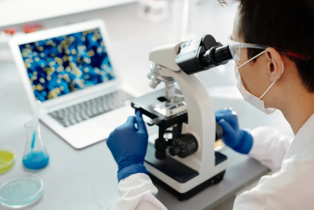 A researcher in gloves and a mask looking through a microscope in a lab