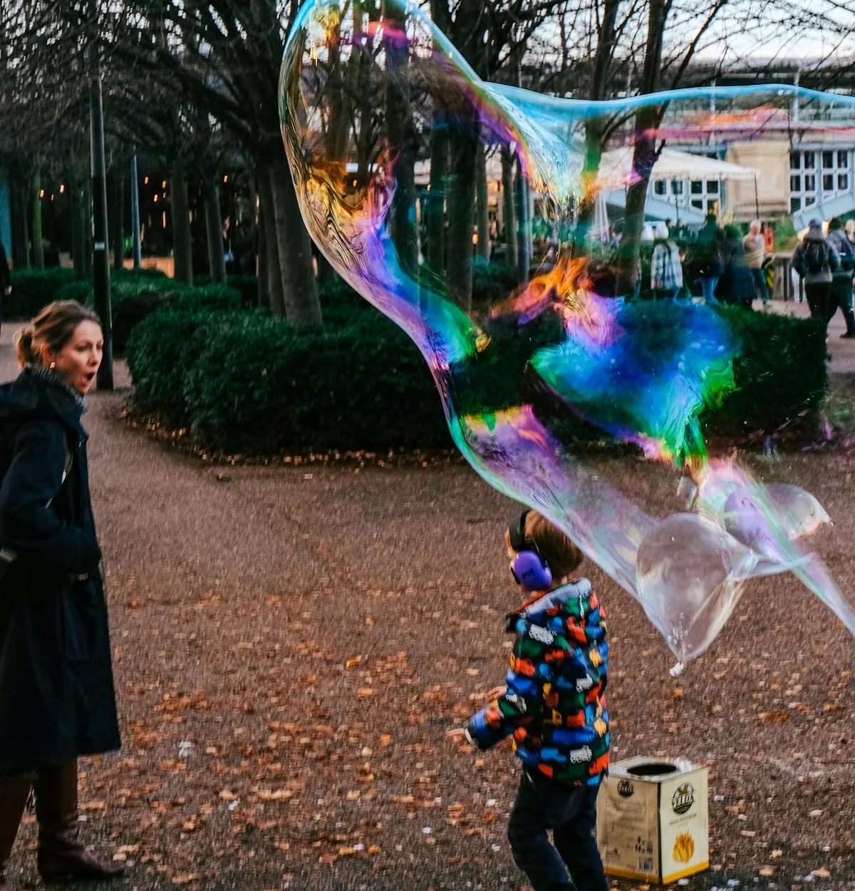 A young boy chasing a giant rainbow bubble in a London park while his mum looks on in delight